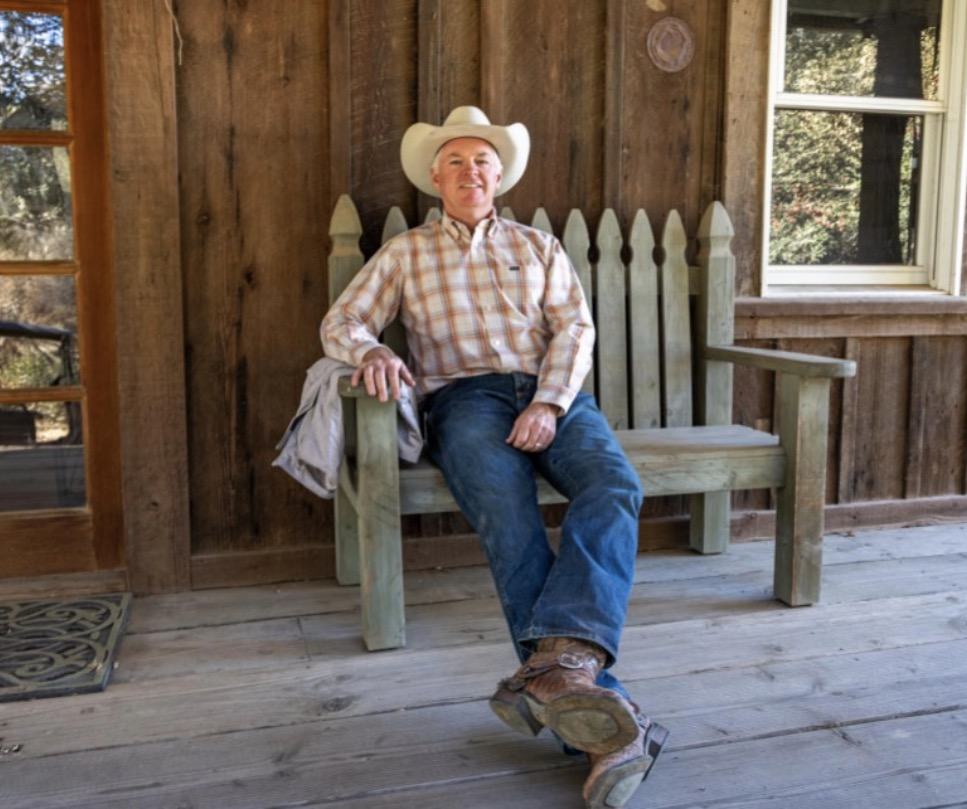 James Ontiveros sitting on a wooden bench on the porch of a ranch cabin, in boots and a cowboy hat.