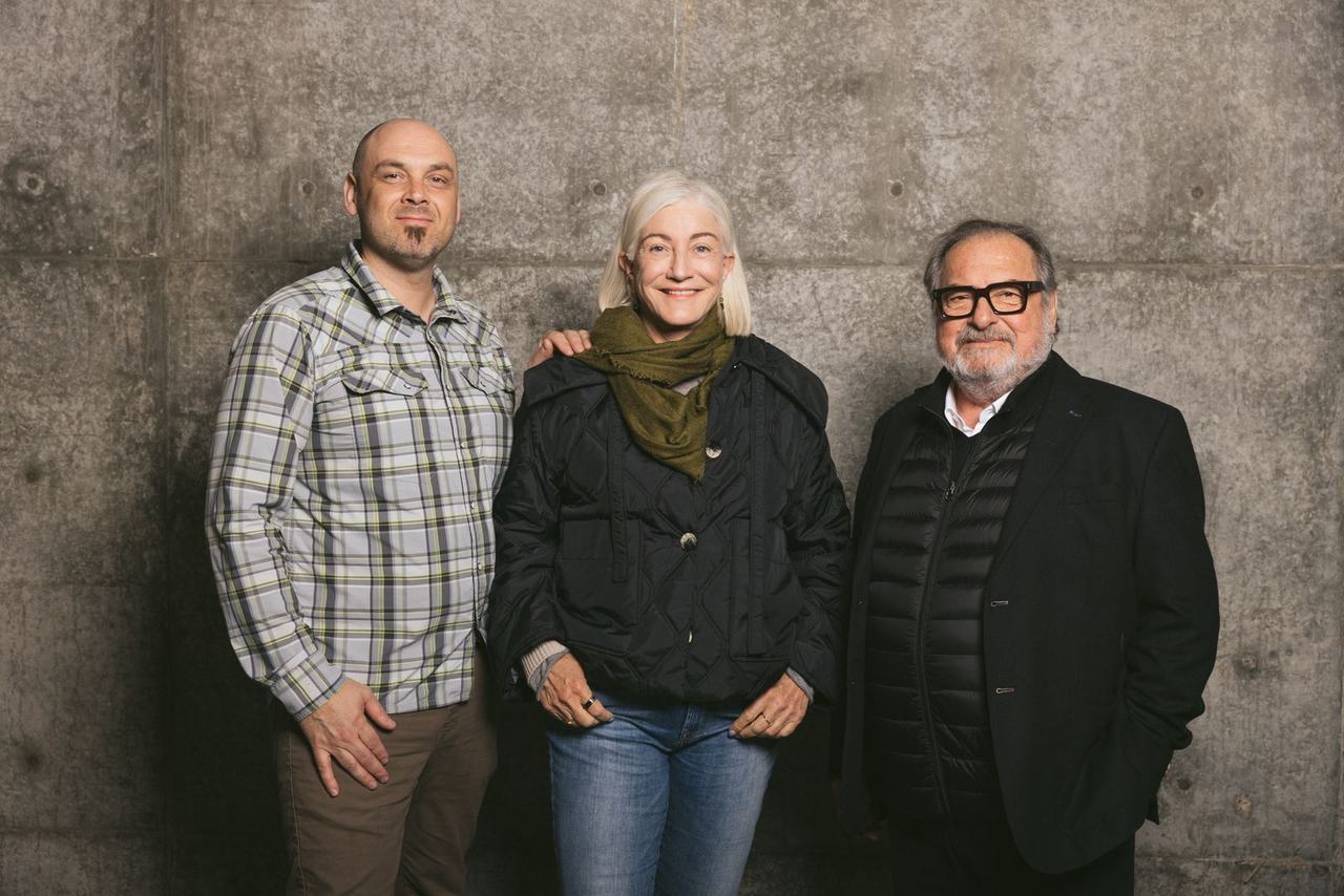 Two men and a woman stand against a concrete wall in the winery.