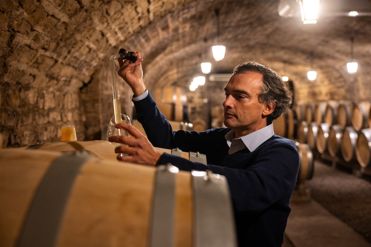 A man holding up a glass pipette of white wine over a barrel in a vaulted cellar