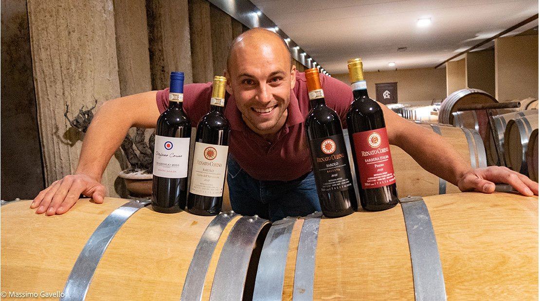 Stefano Corino leaning over a barrel behind four bottles: his Roncaglie, the Rocche, the Riserva and the Barbera Pozzo