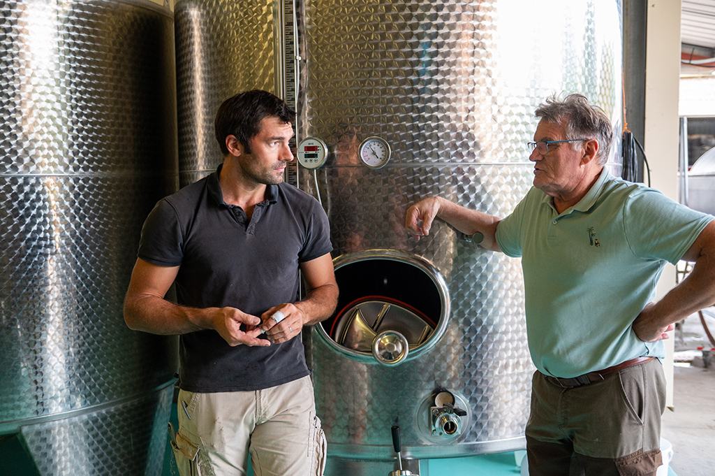 Two men talking beside stainless-steel vats in the cuverie at harvest time.