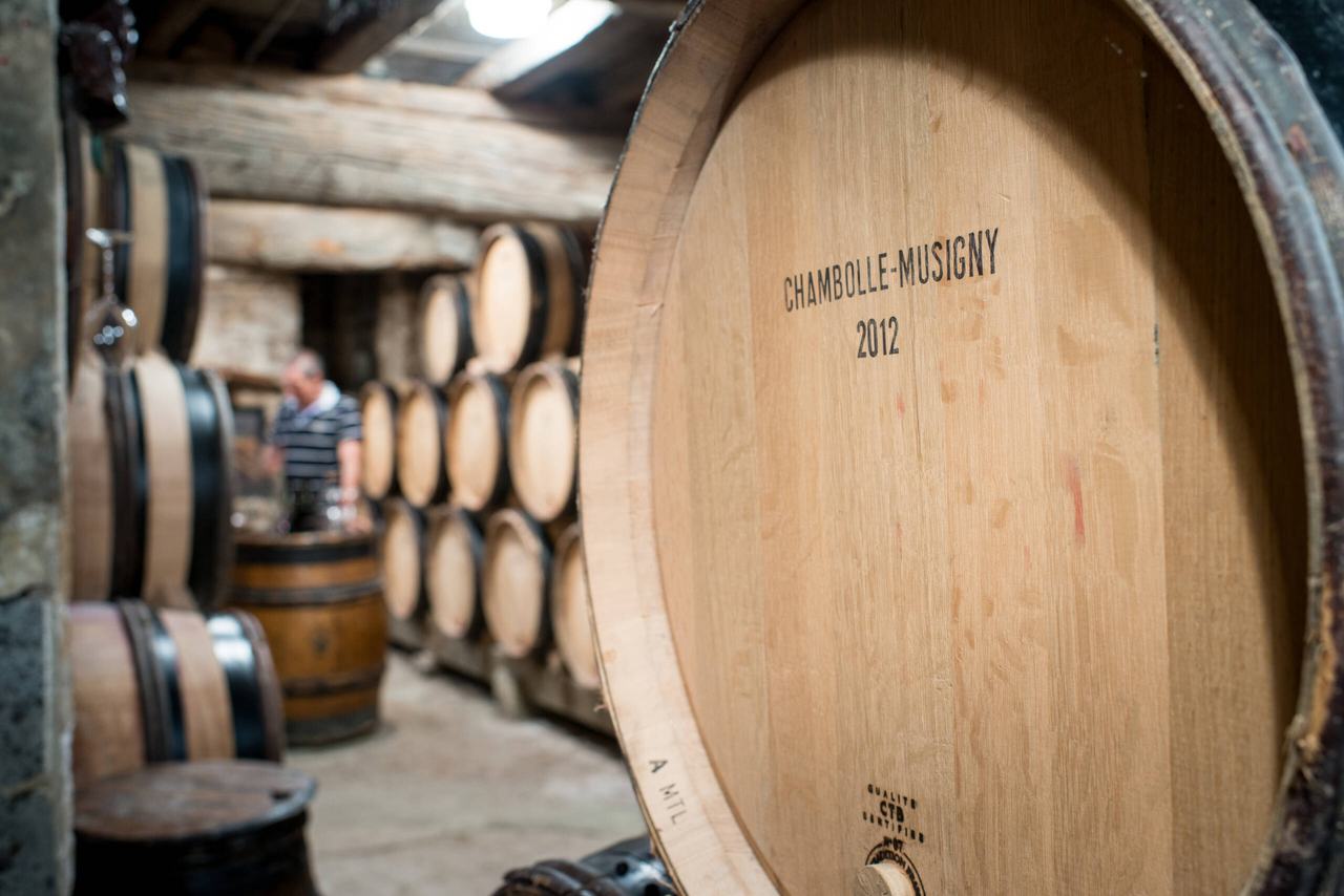 A barrel marked Chambolle-Musigny 2012 in the foreground of a stone cellar lined with casks.