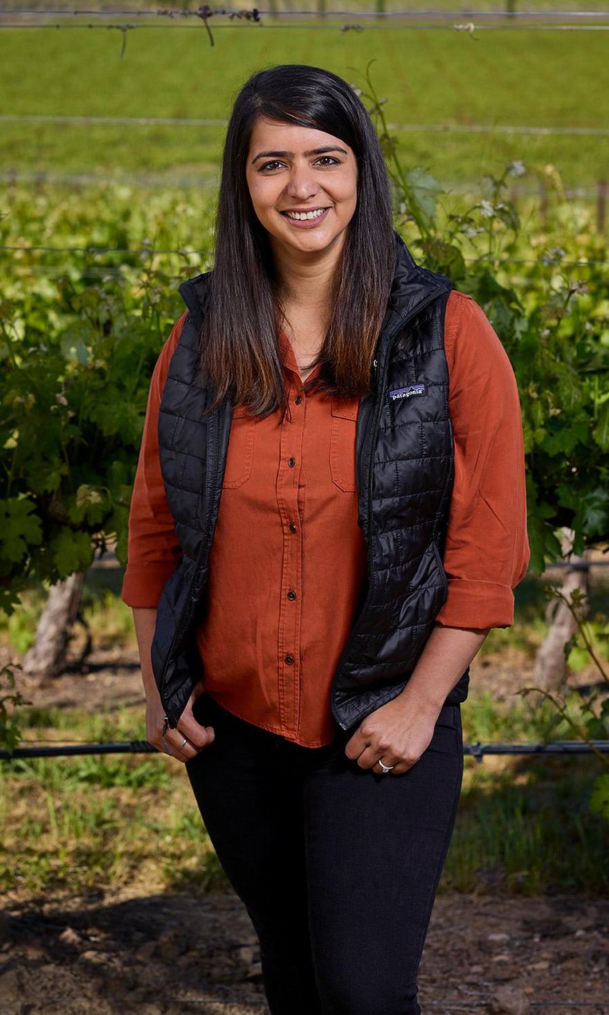 Priyanka French standing in a vineyard in a rust shirt and dark vest.
