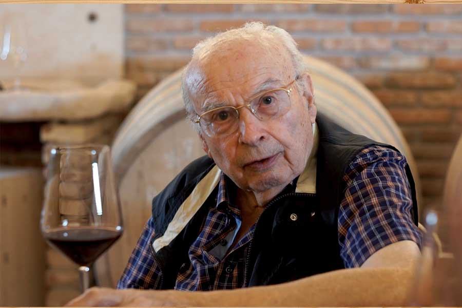 An elderly man in glasses sitting with a glass of red wine in front of a barrel