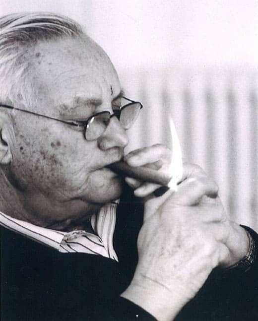 Roger Piquet, an older man in glasses, lighting a cigar