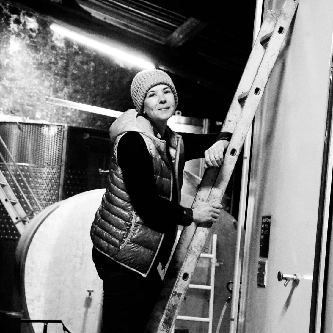 A woman in a knit hat and gilet on a ladder against a concrete tank in the winery, in black and white.