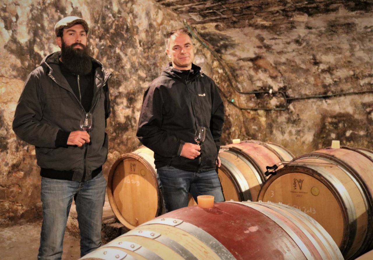 The two partners with glasses of red wine among barrels in the old stone cellar.