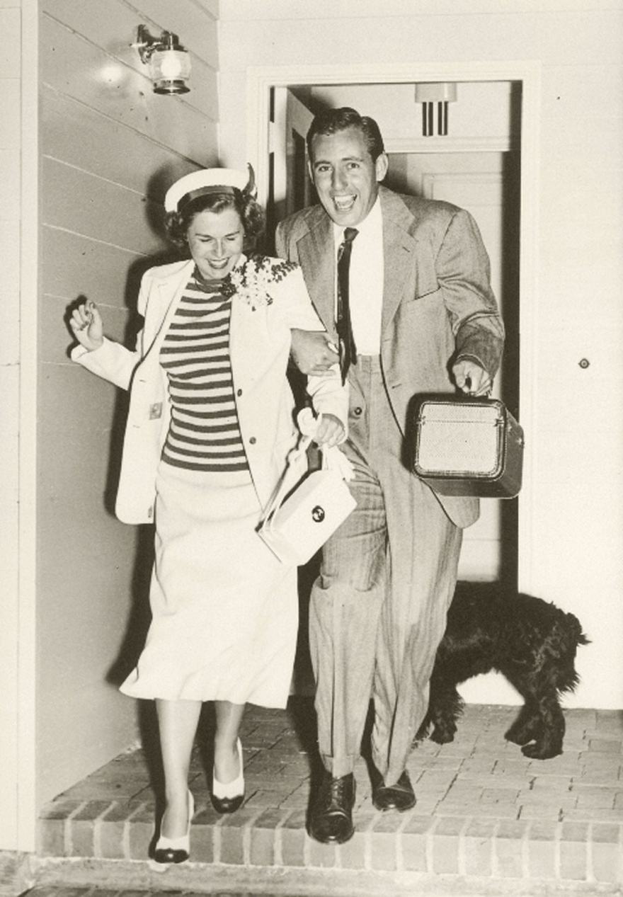 An old black-and-white photo of a smiling couple leaving a house with a dog.