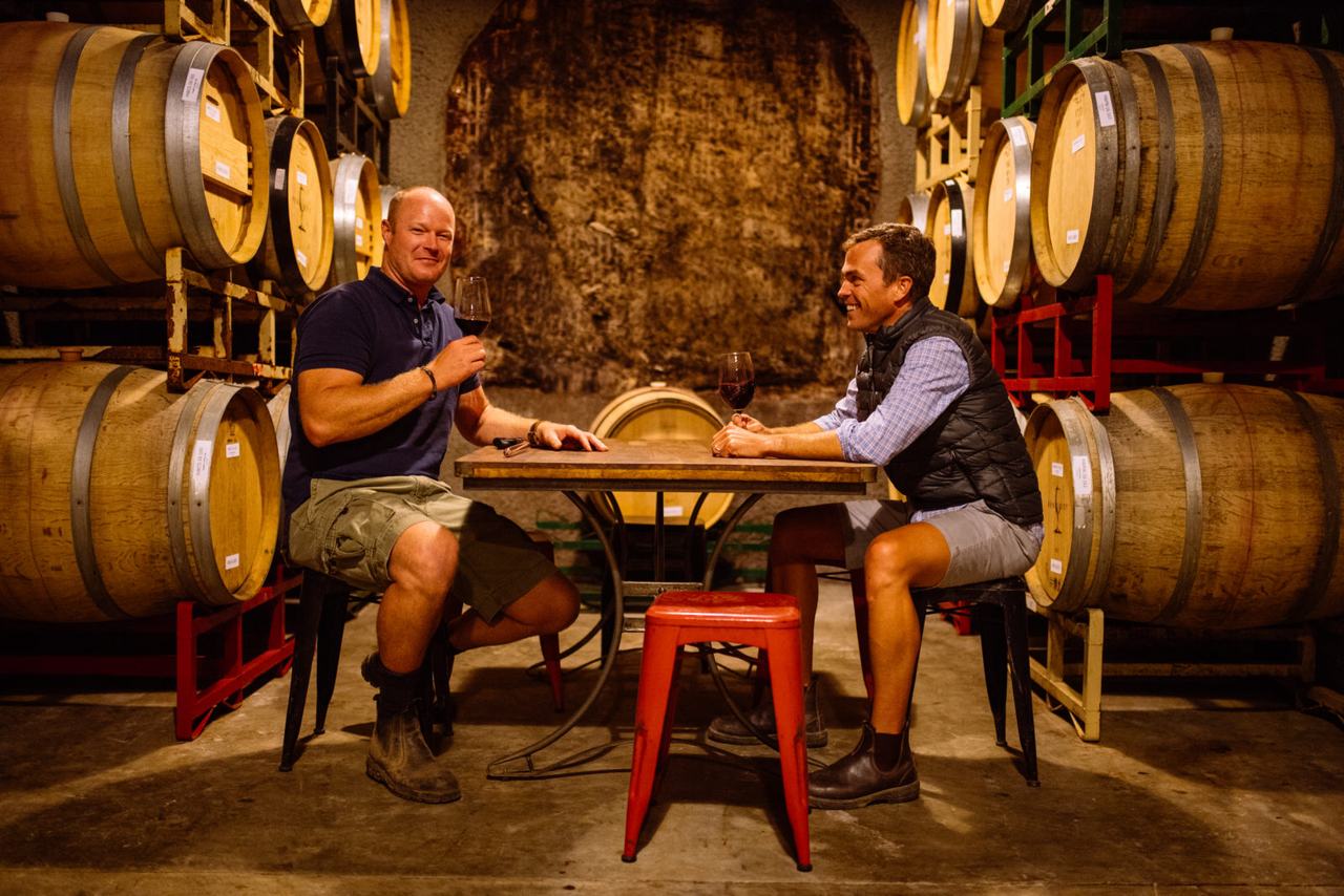 Two men tasting red wine at a small table between stacked barrels in the caves.