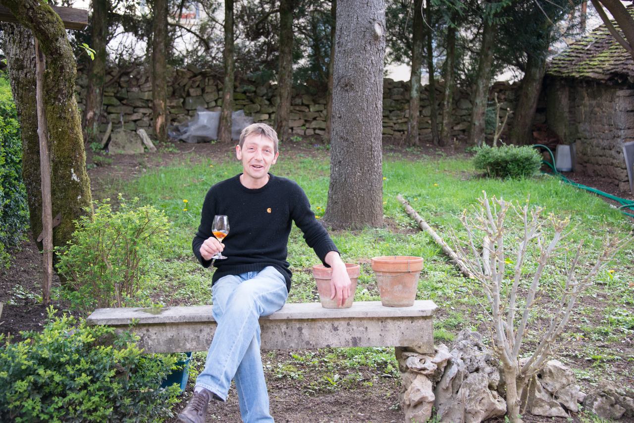 Julien Labet on a stone bench in the garden with a glass of amber Savagnin, a dry-stone wall behind.