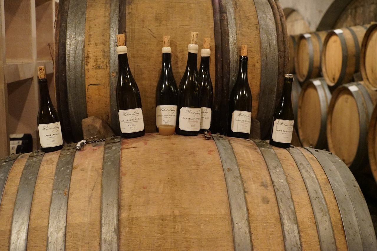 Seven bottles of Hubert Lamy 2013 standing on a barrel in the cellar: La Princée, Derrière chez Edouard Haute Densité, Criots-Bâtard-Montrachet, Santenay Clos des Gravières, Chassagne La Goujonne, Les Frionnes