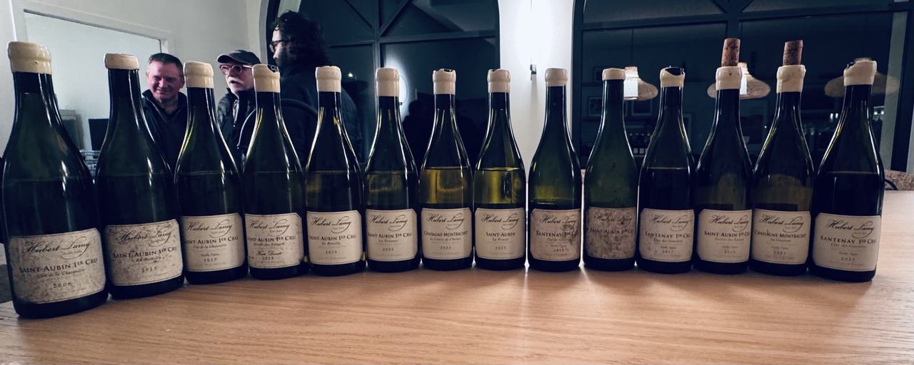 Fourteen bottles of Hubert Lamy lined up on a table, Saint-Aubin premiers crus from 2008 to 2023, Olivier and the tasters reflected in the window behind
