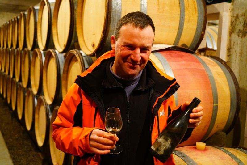 Olivier Lamy smiling in the cellar, a glass in one hand and a bottle in the other, barrels behind