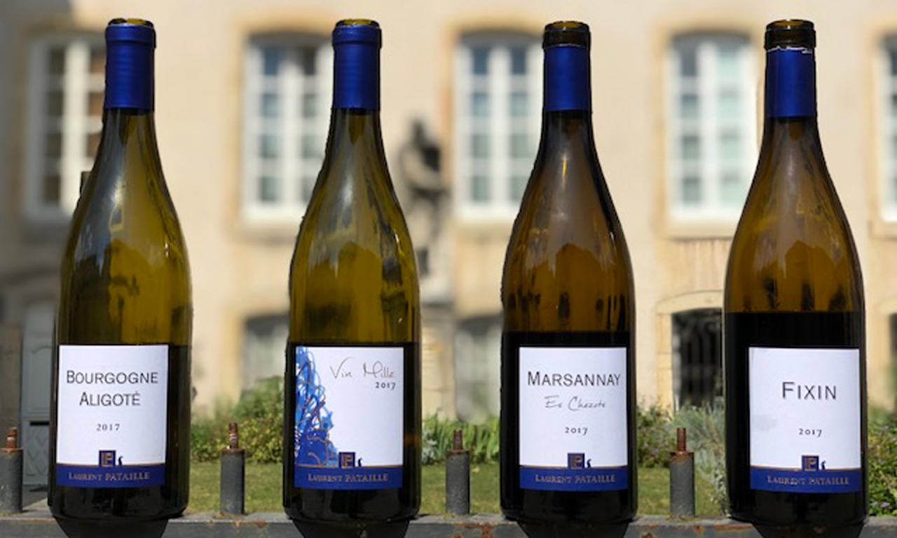 Four bottles of Laurent Pataille 2017 (Aligoté, Vin Mille, Marsannay Es Chezots, Fixin) on a railing in front of a stone building.