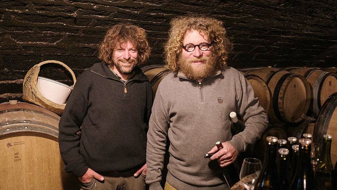 Laurent and Sylvain Pataille side by side in the cellar, bottles on a barrel beside them.