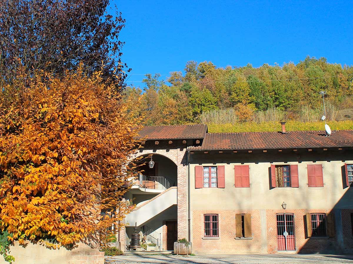 The old Malabaila farmhouse with red shutters in autumn, a tree in orange leaf beside it and woods on the hill behind