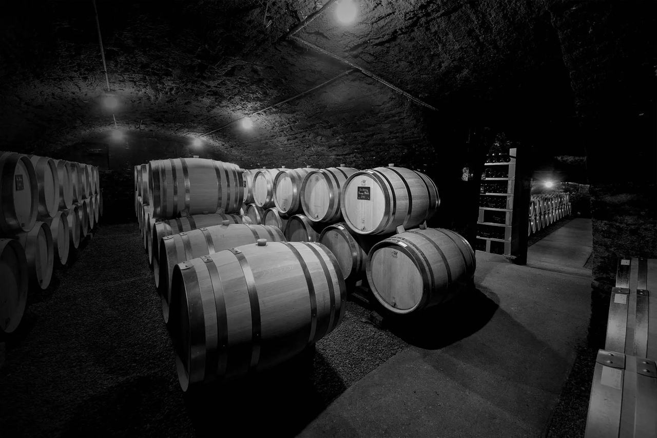 Rows of oak barrels in an arched underground cellar, in black and white.