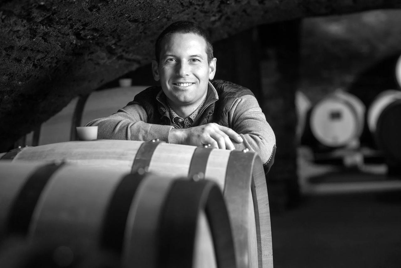 A smiling man leans on a barrel in a vaulted stone cellar, in black and white.