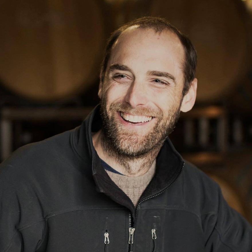 Portrait of winemaker Justin Willett smiling, with barrels behind him.