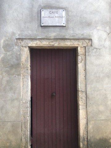 A dark red wooden door in a stone frame in a rendered wall, with a small marble plaque above reading Cave Jean-Marc Pavelot