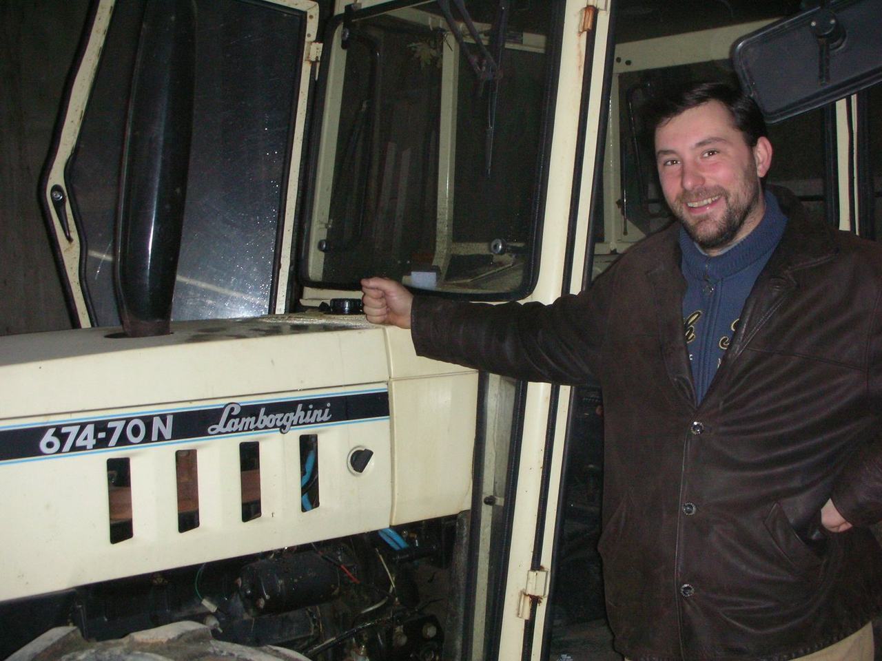 A younger Hugues Pavelot in a brown leather jacket, grinning, one hand on the bonnet of a cream Lamborghini 674-70N tractor in the shed