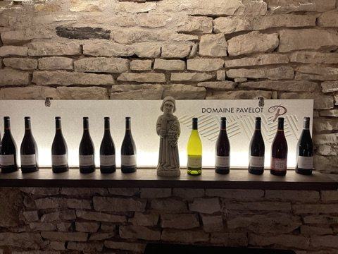 A row of Domaine Pavelot bottles on a lit shelf against a dry-stone wall, a small stone statue of a saint among them, under a Domaine Pavelot banner