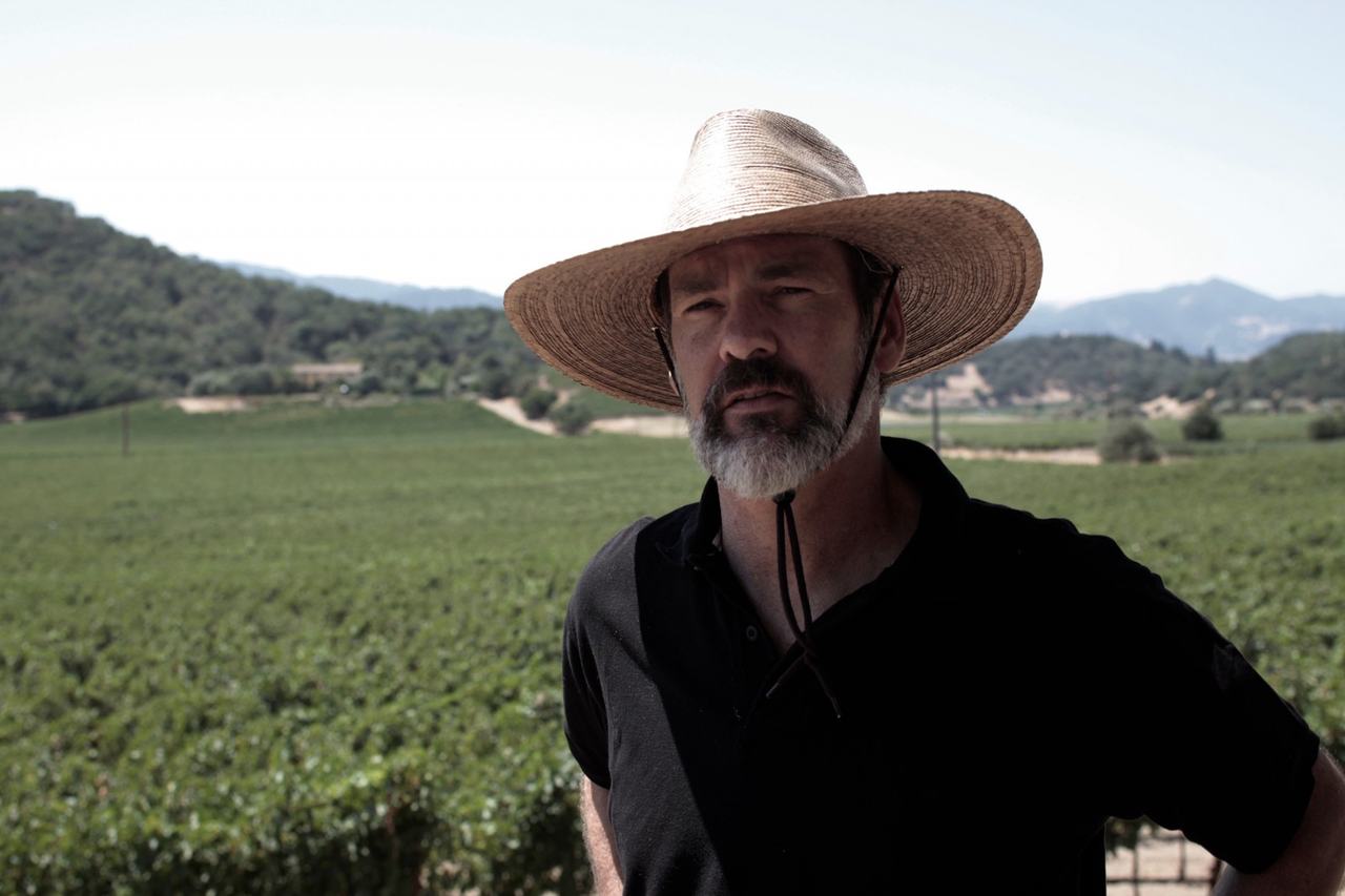 Aaron Pott in a wide straw hat with vineyards and hills behind him.