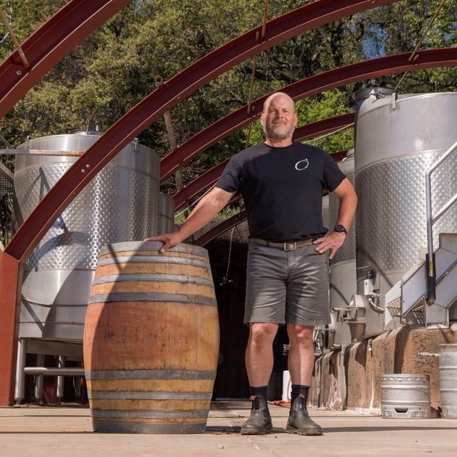 A man in shorts with one hand on a barrel in front of steel tanks
