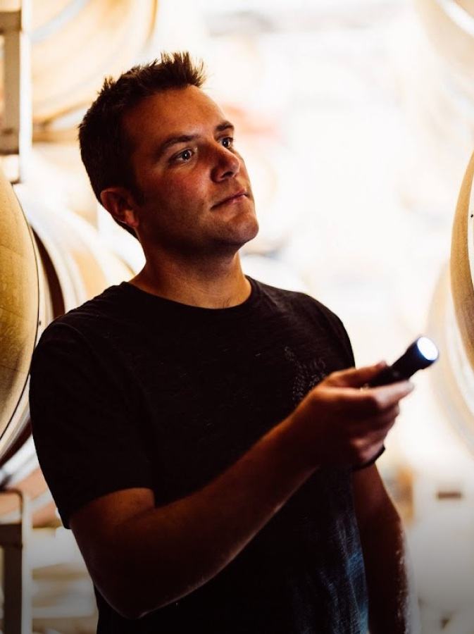 A man in a black T-shirt holds a wine thief among barrels.