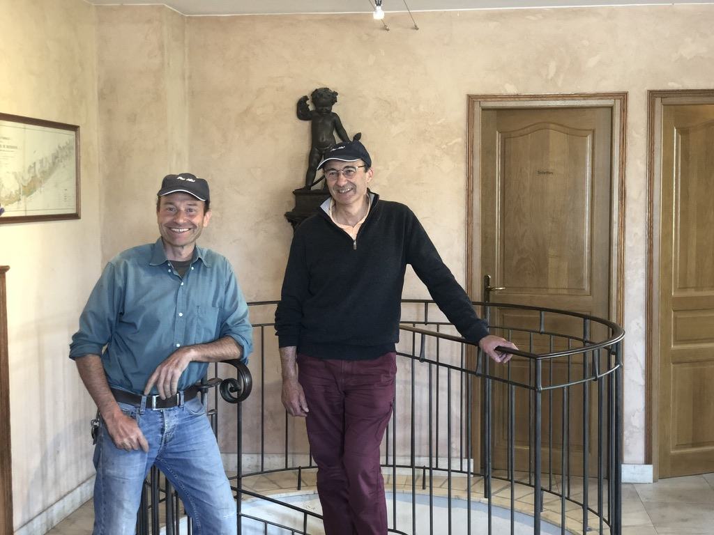 David and Nicolas Rossignol in Pas Mal caps, leaning on the iron rail of the spiral staircase at the domaine, a bronze cherub on the wall behind