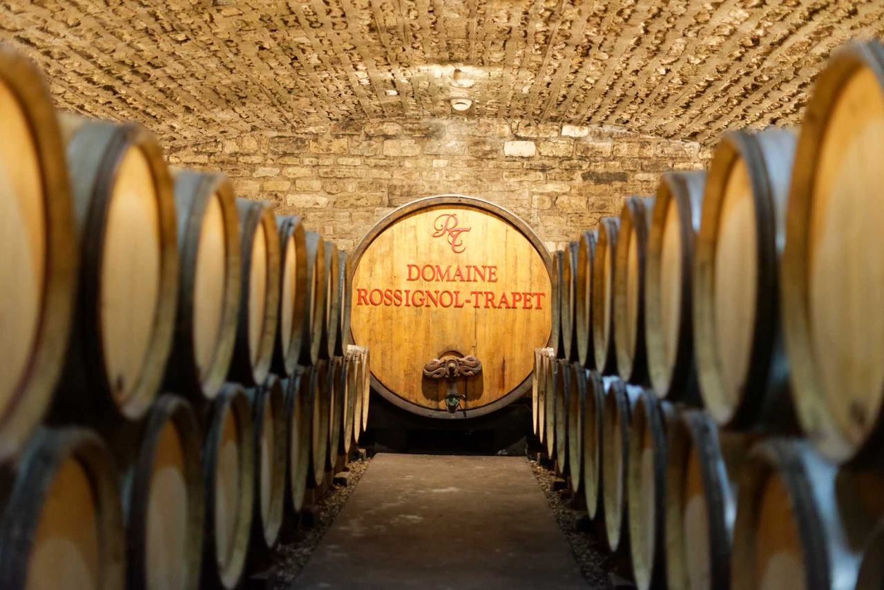 The vaulted stone cellar at Rossignol-Trapet, barrels in two rows leading to a foudre painted Domaine Rossignol-Trapet in red, with the RT monogram