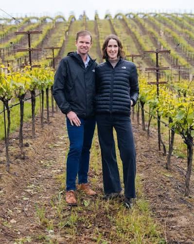 Brian Ball and Laura Jones standing in a vineyard row in spring, mustard in bloom.